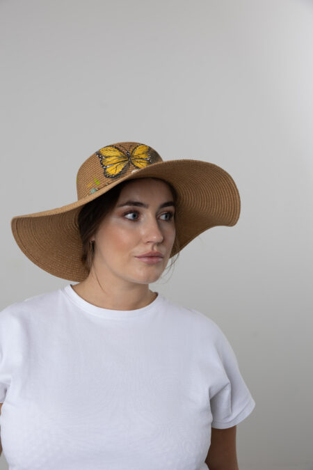 Woman wearing a hand-painted floral straw hat in a sunlit garden or beach.